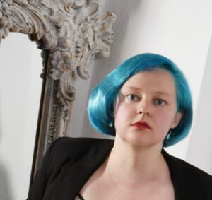 Miss Lia mit blauen Haaren und rotem Lippenstift vor einem Spiegel