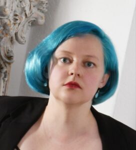 Porträtfoto einer Person mit kinnlangem, leuchtend türkisblauem Haar, hellem Teint und rotem Lippenstift. Sie blickt direkt in die Kamera. Der Hintergrund ist hell und ornamental.